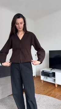 Frau richtet eine dunkelbraune Jacke mit Knöpfen, steht in einem Wohnzimmer mit Holzboden, weißem Fernsehmöbel und einem Flachbildfernseher.