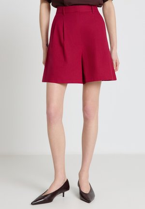 Donna che indossa pantaloncini rossi plissettati a vita alta, lunghezza al ginocchio, abbinati a scarpe marrone scuro con tacco basso e punta affusolata, su sfondo bianco.