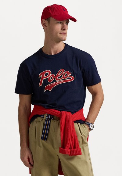 Navy t-shirt with red "Polo" logo, paired with beige trousers and a red sweater tied at the waist, and a red cap.