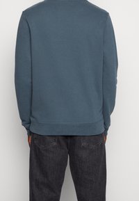 Niebieska bawełniana bluza z długimi rękawami, okrągłym dekoltem, prążkowanymi mankietami i ściągaczem. Noszona z ciemnoszarymi dżinsami, prezentująca prosty, codzienny styl.