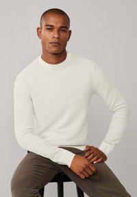 Maglione lavorato a maglia color crema con scollo rotondo, polsini a coste e un motivo testurizzato, abbinato a pantaloni marrone scuro, seduto su uno sgabello nero.