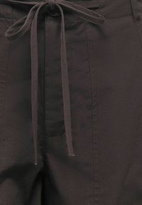 Gros plan d’un pantalon marron foncé avec un cordon en tissu noué à la taille et des détails de couture verticale visibles.