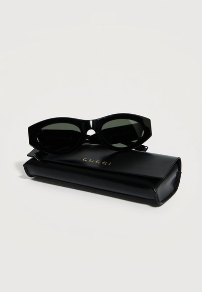 Γυαλιά ηλίου με μαύρους φακούς σε σχήμα γάτας, τοποθετημένα πάνω σε μαύρη θήκη Gucci με χρυσά γράμματα σε λευκό φόντο.