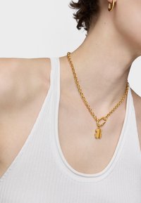 Collier en chaîne dorée avec un pendentif en forme de verrou texturé, porté sur un débardeur blanc à côtes, avec un décolleté rond.