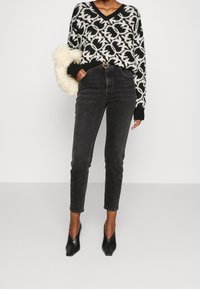 Person i en svart och vit mönstrad tröja, högmidjade svarta jeans med bälte, svarta spetsiga stövlar, håller en handväska i krämfärgad faux päls.