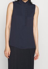 Námořnická bezramínková blůza s vertikálními pruhy, s volánkovým límcem a knoflíkovou légou. Tkanina vypadá hladce s mírným leskem.