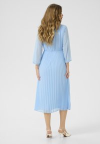 Svijetloplava plisirana haljina s rukavima do tri četvrtine, naglašenim strukom i lepršavim dizajnom, uparen s cipelicama na visoku petu s otvorenim prstima.