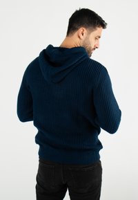 Hoodie en maille bleu marine avec une finition texturée, doté d'une grande capuche et de poignets et ourlet côtelés. Porté avec un jean noir.