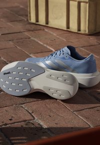Jasnoniebieski but sportowy Adidas z białą podeszwą pokazującą bieżnik Continental, umieszczony na czerwonawym ceglanym podłożu z beżową doniczką w tle.