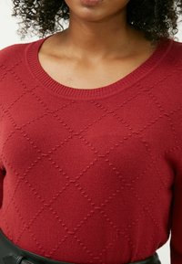 Roter, gestrickter Pullover mit Rautenmuster, geripptem Kragen und strukturierter Oberfläche. Getragen mit einem schwarzen Gürtel, der eine glatte Stoffoberfläche aufweist.