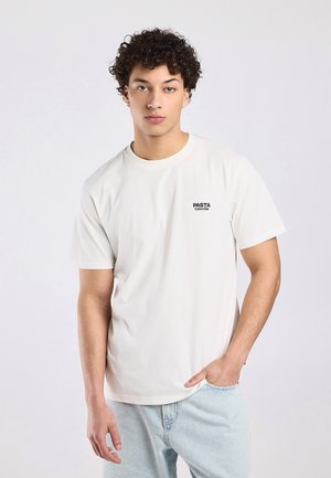 Young man with curly hair wearing a white "PASTA Culture Club" t-shirt and light blue jeans, standing against a plain light background.