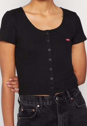Femme portant un crop top noir côtelé à manches courtes avec des boutons et le logo rouge de Levi's, ainsi qu'un jean denim noir taille haute.