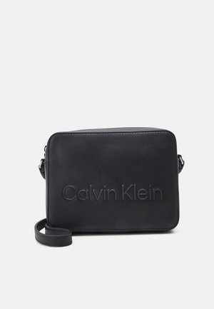 Sac bandoulière rectangulaire noir avec logo Calvin Klein embossé et sangle ajustable sur un fond uni.