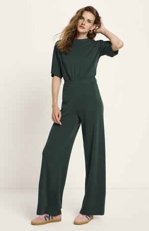 Woman wearing dark green relaxed-fit jumpsuit and pink sneakers, standing with one hand on head against plain background.