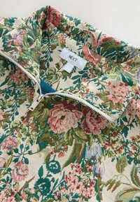 Haut zippé à motifs floraux avec un fond crème orné de motifs floraux verts, roses et bleus, texture tissée et étiquette de marque.