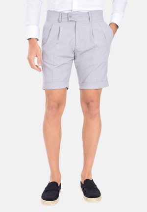 Pantaloni corti grigi chiari su misura con pieghe, orlo risvoltato e chiusura a bottoni sul fronte, realizzati in un tessuto liscio. Indossati con scarpe navy slip-on.