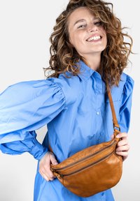 Borsa a tracolla in pelle marrone con forma curva, caratterizzata da un dettaglio con cerniera. La borsa è indossata sopra una camicia blu con maniche a sbuffo.