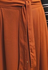 Rustfarvet nederdel med en bundet talje detalje, lavet af glat stof med en blød tekstur og plisseret design, der viser en knaplukning.