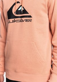 Huppari persikanvärinen, valmistettu pehmeästä kankaasta, jossa on etupuolella kengurutasku, ribbikudotutu cuffit ja suuri musta Quiksilver-logo.