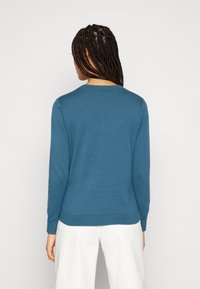 Blauer Pullover mit langen Ärmeln, rundem Ausschnitt und geripptem Saum. Hergestellt aus weichem Stoff mit glatter Textur. Weiße Hose ist sichtbar.