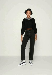 Sweater cropped preto com decote em pedras, combinado com jeans pretos de cintura alta e sapatilhas pretas. A modelo usa um chapéu preto.
