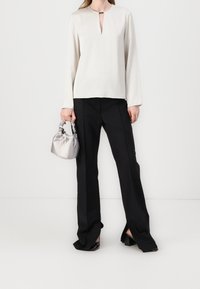 Haut crème à manches longues avec une encolure goutte d'eau, associé à un pantalon noir à jambes larges. Le mannequin tient un petit sac argenté. Tissu lisse, design minimaliste.