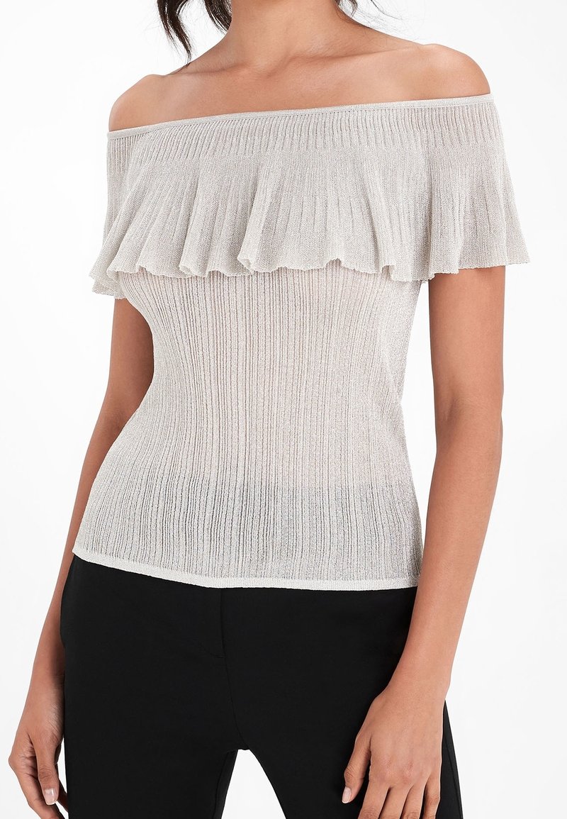 Femme portant un haut en maille côtelée ajusté, épaules dénudées et col à volants en beige clair, assorti à un pantalon noir. Fond blanc.
