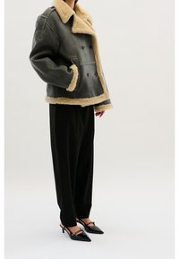 Chaqueta de borrego gris con un amplio cuello beige, cierre cruzado y bolsillos laterales, combinada con pantalones negros y tacones puntiagudos negros.