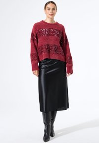 Vrouw in een rode sweater met sequins, zwarte midi-rok en zwarte kniehoge laarzen, staande tegen een effen witte achtergrond.