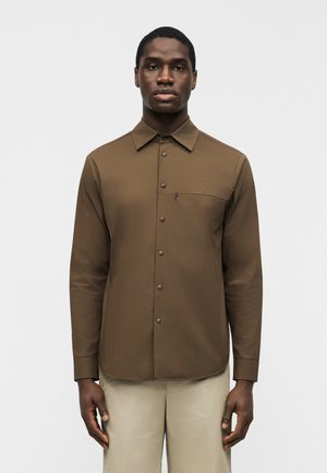 Egy férfi, aki hosszú ujjú, barna, gombos ingben áll, mellkasi zsebbel és világos bézs nadrágot visel, egy sima fehér háttér előtt.