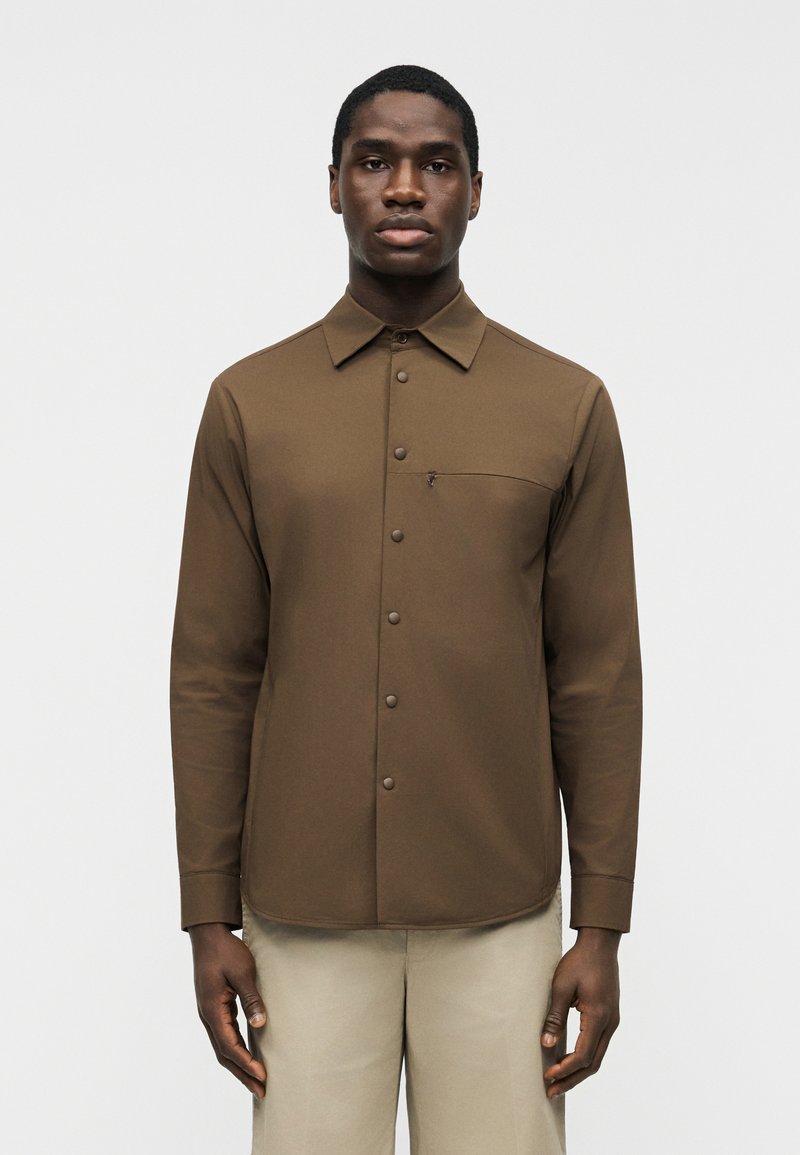 Άνδρας που φορά καφέ πουκάμισο με μακρύ μανίκι, κουμπιά και τσέπη στο στήθος, καθώς και ανοιχτό μπεζ παντελόνι, στέκεται μπροστά σε απλό λευκό φόντο.