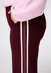 Bordeauxkleurige broek heeft een zijstriping in zwart, rood en crème. De stof lijkt rekbaar, met een relaxte pasvorm en zijzakken.