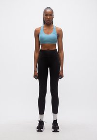 Γυναίκα με μπλε αθλητικό σουτιέν και μαύρο κολάν κρατώντας αλτήρες 2 λιβρών στα πλευρά της, στέκεται μπροστά σε απλό λευκό φόντο.