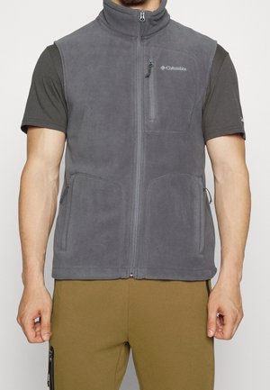 Muž stojící na jednoduchém pozadí, oblečený v šedé fleecové vestě Columbia na zip přes tmavé tričko s krátkým rukávem a hořčicové kalhoty.