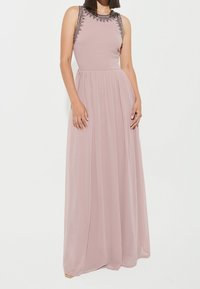 Robe longue sans manches en rose pâle, avec un col orné de perles, une jupe fluide et un tissu léger avec une texture douce.