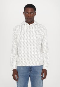 Vit hoodie med ett geometriskt grått mönster, dragsnören och ribbade ärmmuddar, ihop med blå jeans. Mjuk textur, avslappnad design.