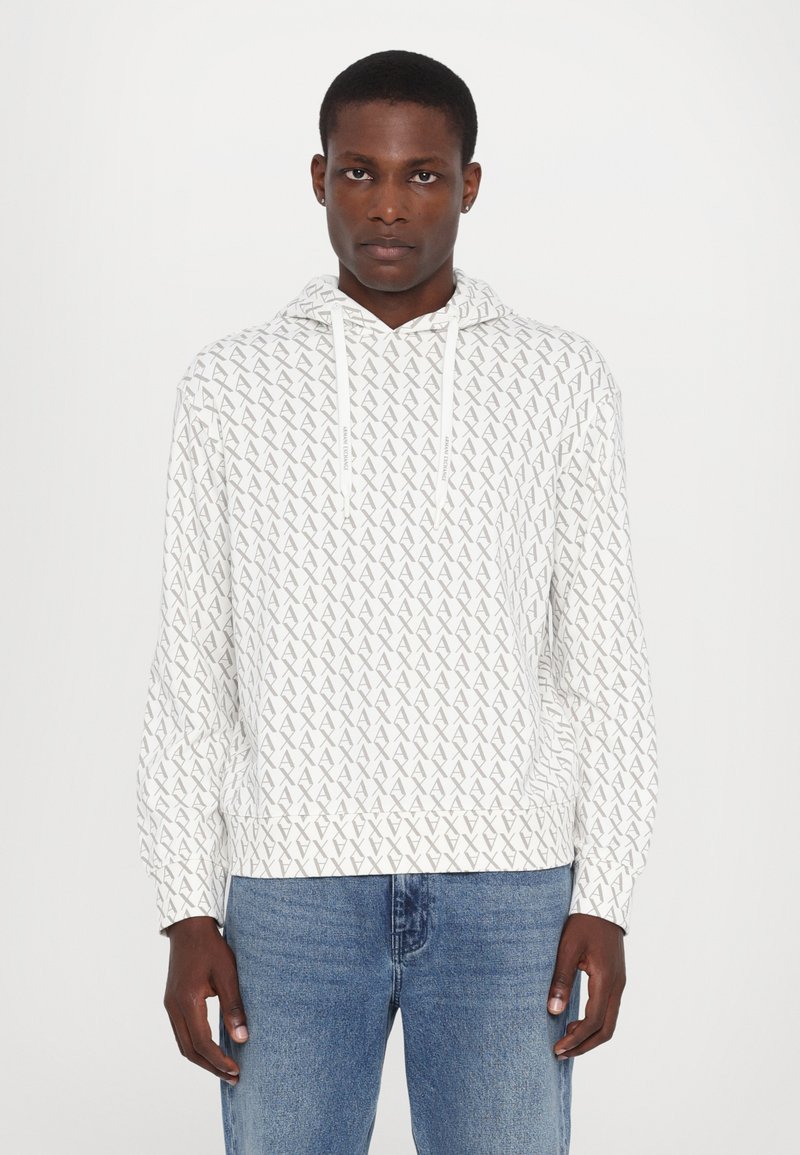 Vit hoodie med ett geometriskt grått mönster, dragsnören och ribbade ärmmuddar, ihop med blå jeans. Mjuk textur, avslappnad design.
