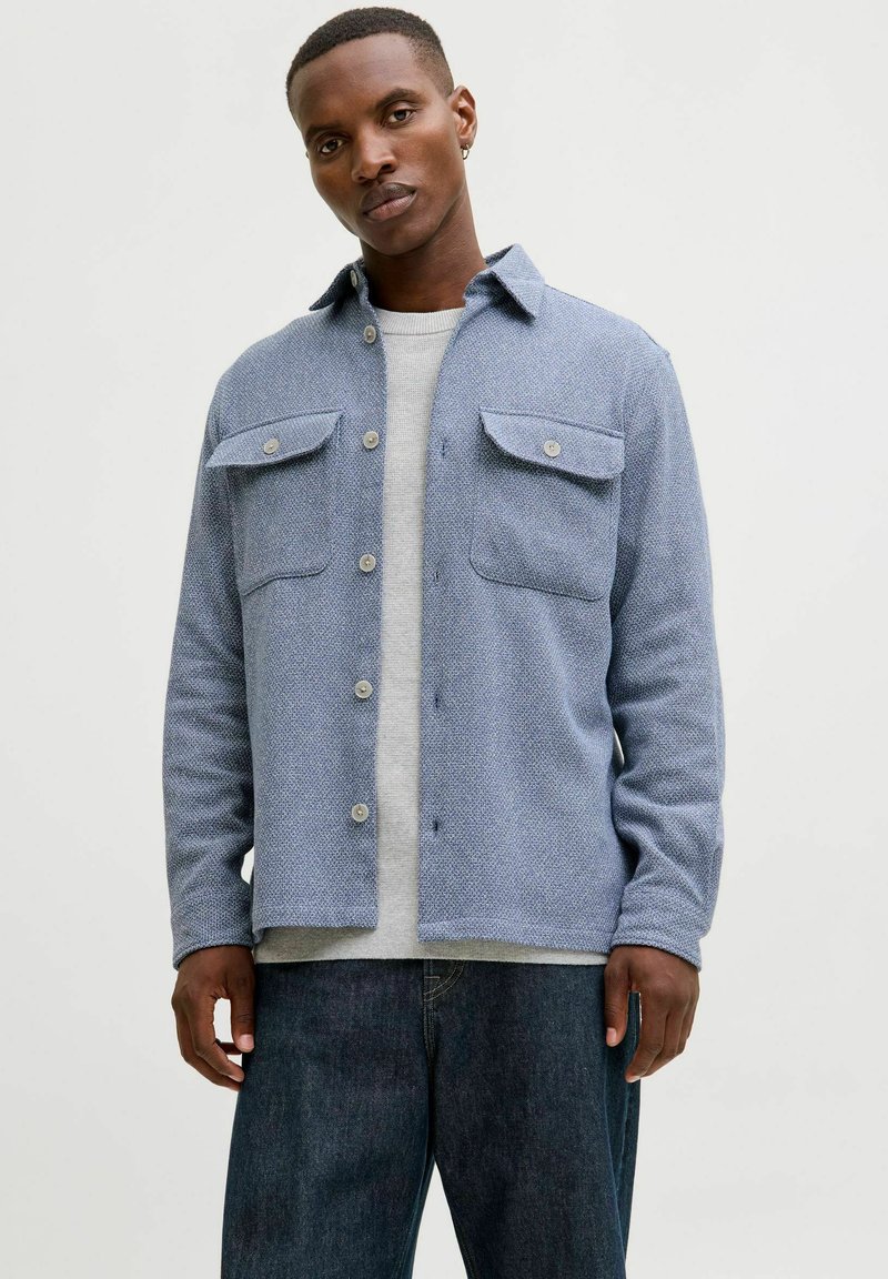 Blå teksturert skjorte med knappelukking og to brystlommer, kombinert med en grå genser under og mørke denimjeans. Casual design.