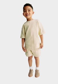 Light beige knit short-sleeve shirt and matching shorts. Worn with tan sneakers and white socks. Smooth texture, relaxed fit, minimal seams.