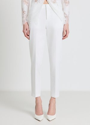 Pantaloni bianchi dritti su misura indossati con décolleté bianche appuntite e un top bianco in pizzo a maniche lunghe, in piedi davanti a uno sfondo bianco.