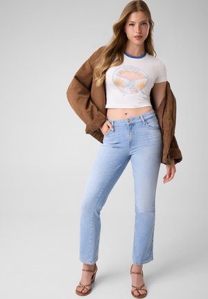 Frau trägt hellblaue Jeans, ein weißes Cropped-Grafik-T-Shirt, eine braune offene Jacke und braune Sandalen mit Absatz, steht mit einer Hand in der Tasche.