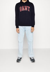 Sweat à capuche bleu marine avec lettrage rouge "GANT", associé à un jean bleu clair et des baskets noires. Coupe décontractée, texture de tissu douce.