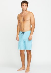 Shorts de bain bleu clair en matériau lisse, avec un cordon de serrage noir, un emblème sur la jambe gauche et une coupe décontractée.