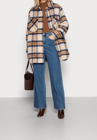 Beige rutigt fodral med blå och orange linjer över en brun polo, tillsammans med högmidjade blå jeans och bruna klackstövlar.