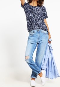 Marinblommig kortärmad topp, ljusblå slitna jeans med ett rivet knä, och vita slip-on skor; ljusblå skjorta hängande över armen.