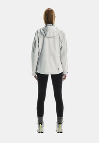 Ljusgrå vattentät jacka med huva, lös passform och elastiska ärmslut, ihop med svarta leggings och sneakers med ljusgula accenter.