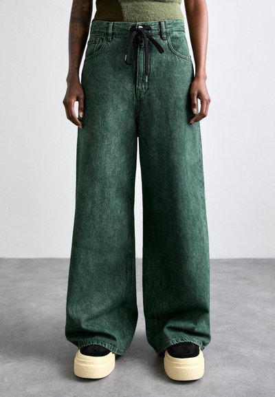 Pantaloni cu picioare largi din denim verde închis, având un brâu cu șnur și o textură subtilă. Asortați cu adidași negri și talpă crem.