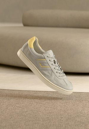 Pantof sport gri deschis din piele întoarsă perforată, cu un accent galben pe călcâi, șireturi plate și talpă albă din cauciuc.