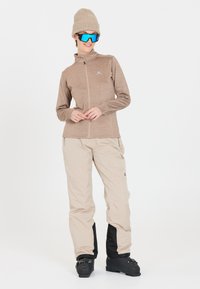 Beige zip-jakke med tekstureret finish, parret med beige skibukser med sorte accenter og sorte skistøvler. Blå solbriller og en strikket hue.