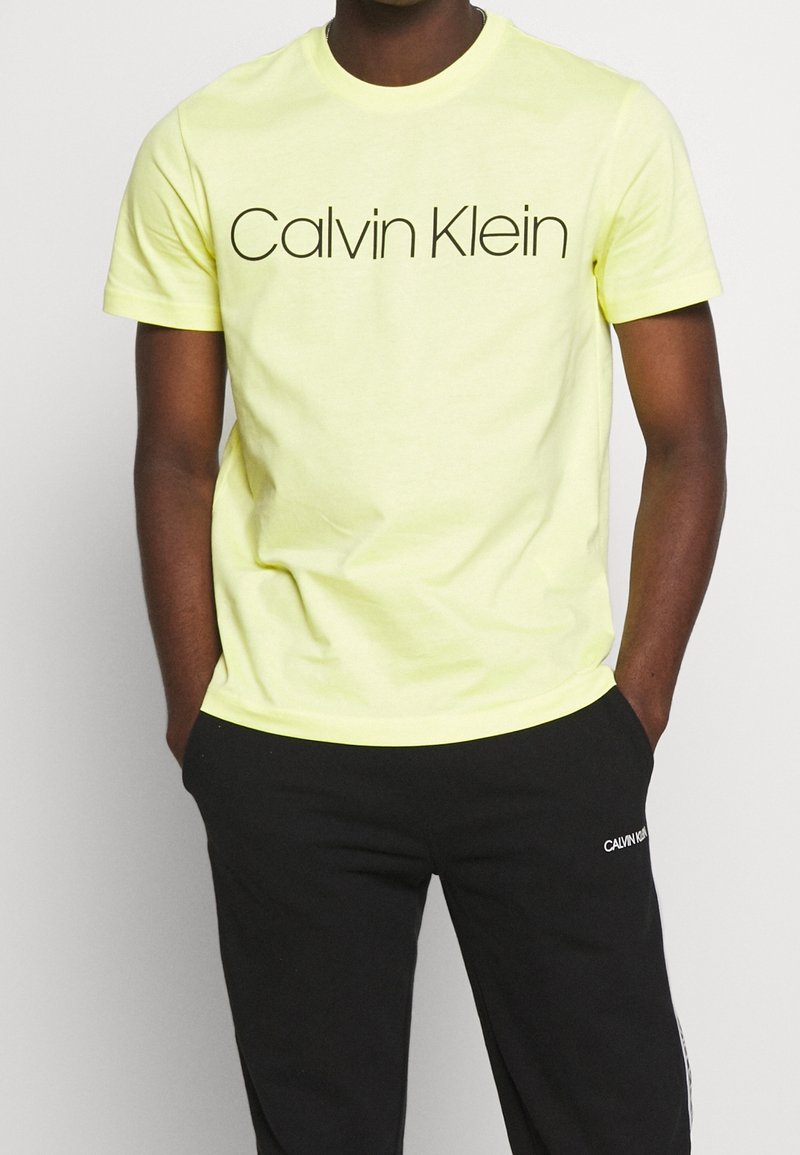 Bright yellow cotton t-shirt featuring black "Calvin Klein" logo on the front. Short sleeves, crew neck, and a relaxed fit. Paired with black pants.